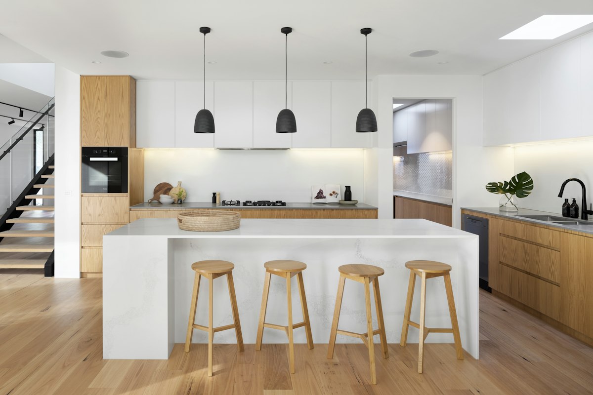 Modern open-concept kitchen featuring a white marble waterfall island, 4 wooden stools, light wood floors, and natural oak cabinets.